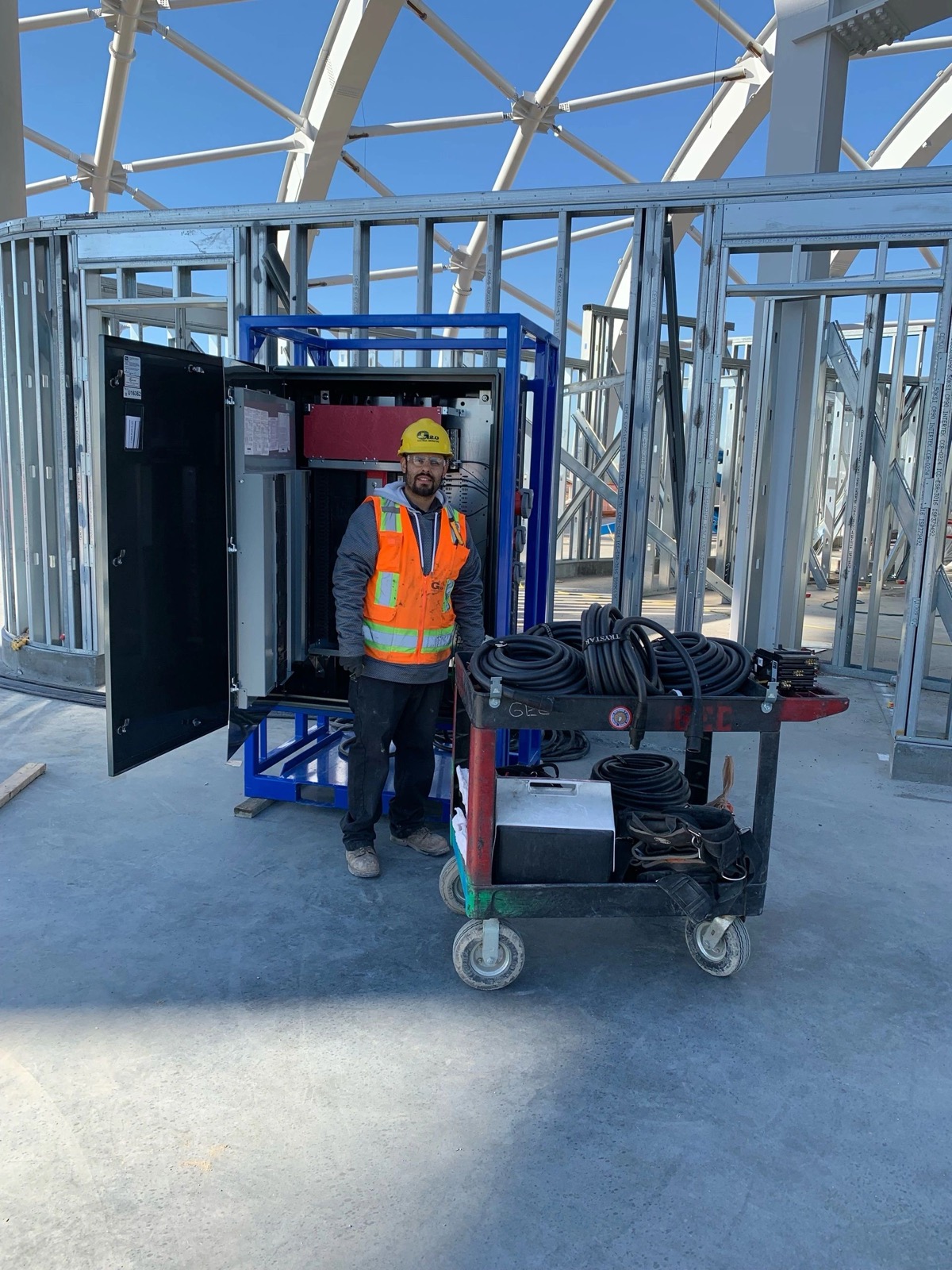 A G2.0 electrician in a branded hard hat, safety glasses and high visibility vest standing at an open switchgear enclosure inside a steel framed structure, a cart of coiled cable beside him.