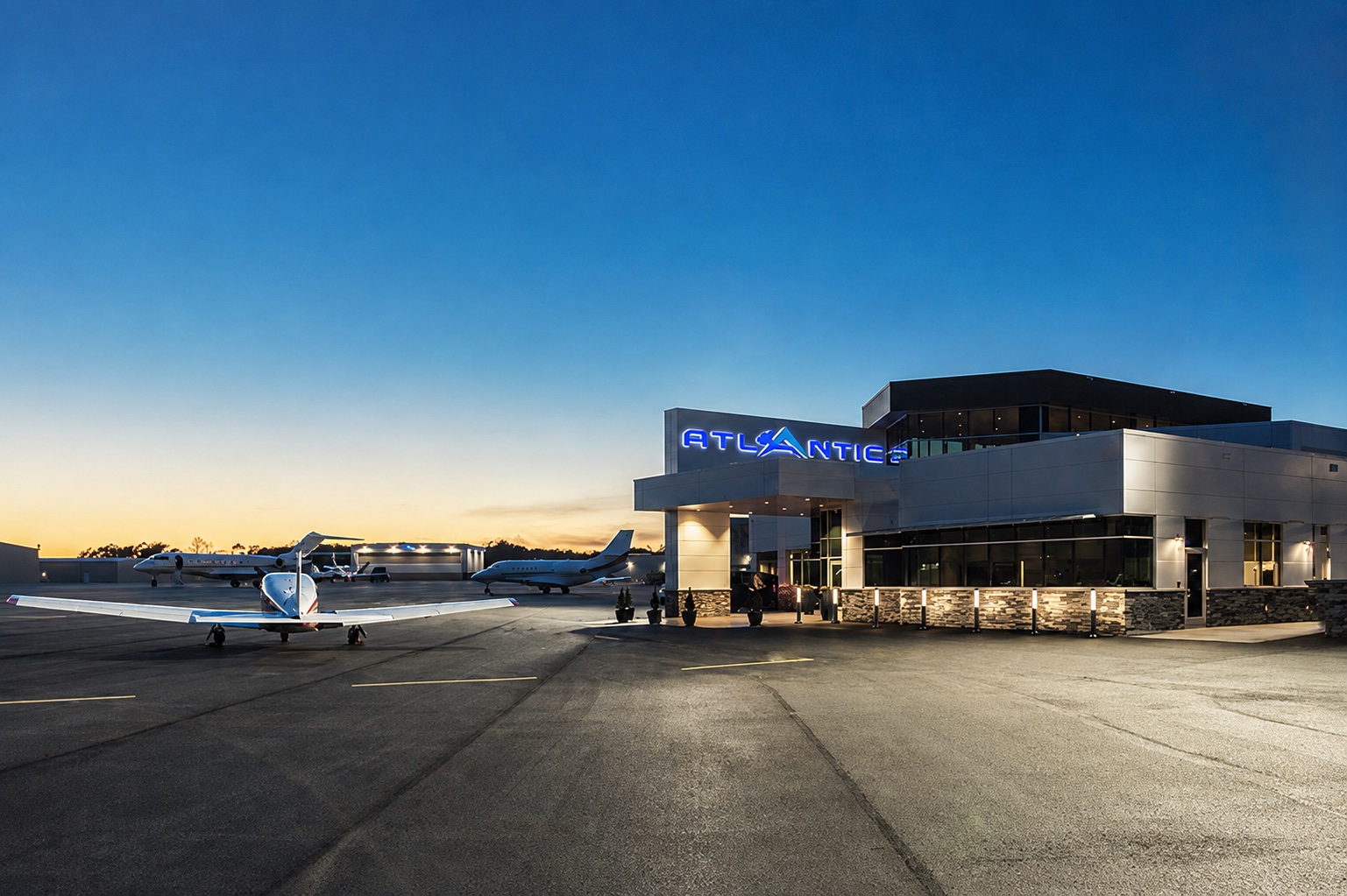 An Atlantic Aviation fixed base operator terminal at dusk, the lit ATLANTIC sign above the entrance canopy and business jets parked on the apron beyond.