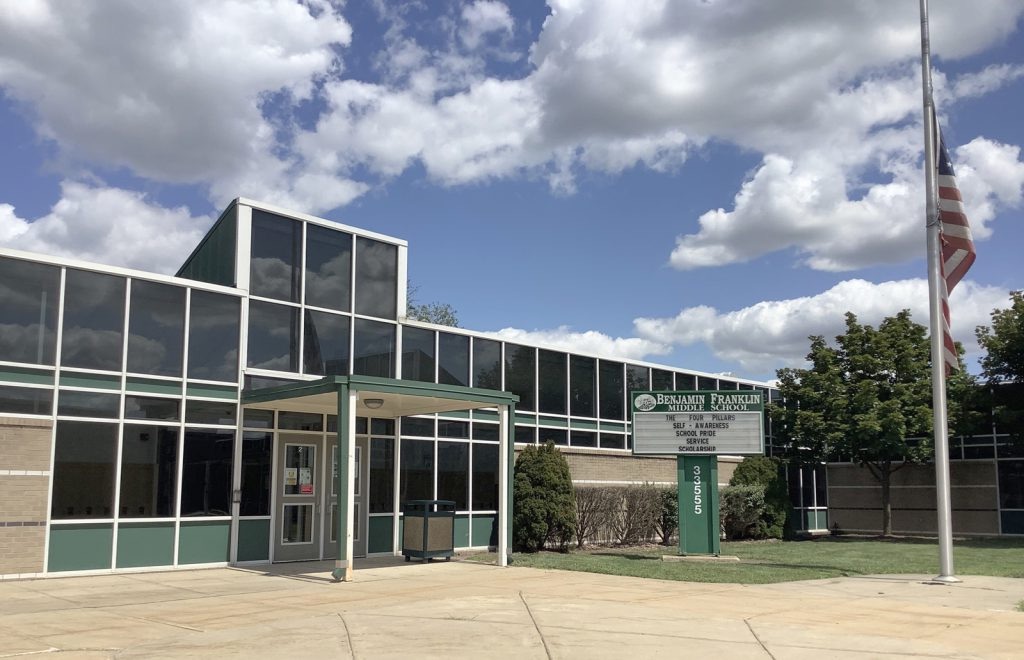 The entrance to Benjamin Franklin Middle School, a low glazed frontage with green trim, a marquee sign and a flagpole.