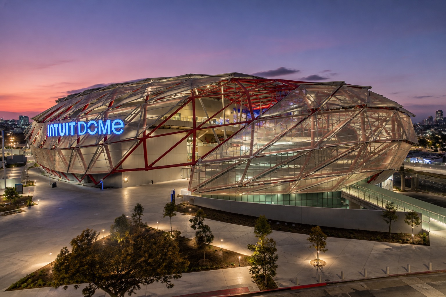 Intuit Dome at dusk, its faceted lattice shell lit from within over the plaza, the name in blue across the west elevation.