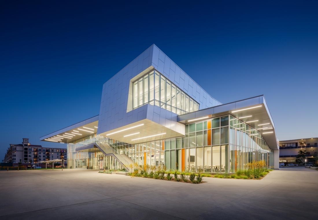 A college building at dusk, a white clad upper volume cantilevered over a glazed ground floor with an external stair and a lit soffit.