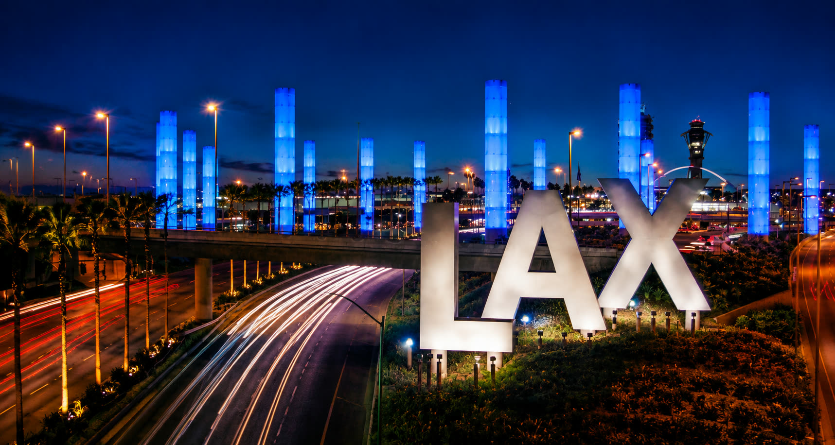The LAX gateway pylons lit blue at dusk behind the airport's letter sculptures, with traffic streaking along Century Boulevard.