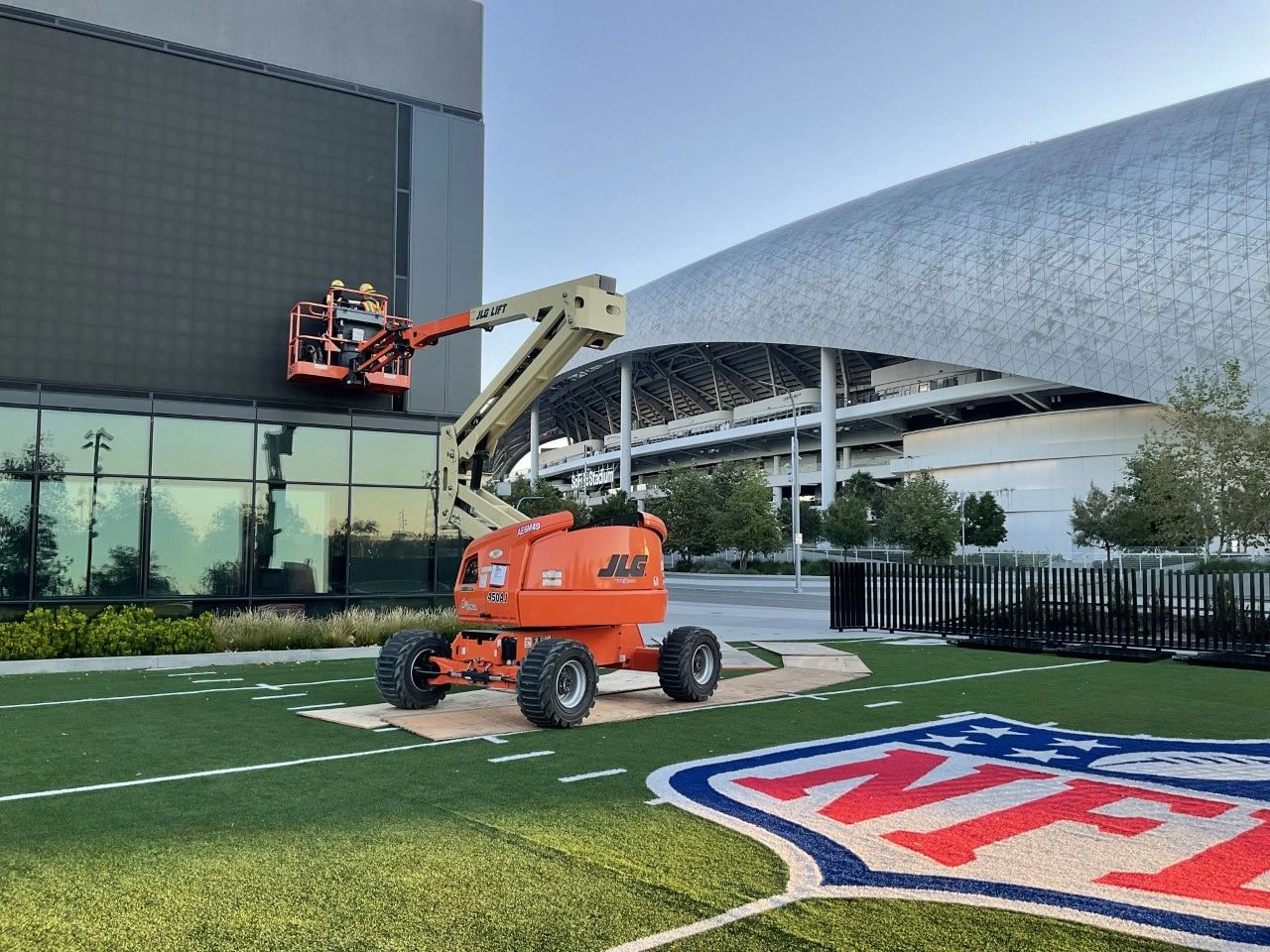 Two G2.0 electricians working from a raised boom lift on the facade of the NFL building at Hollywood Park, with the roof canopy of SoFi Stadium behind them and an NFL shield painted on the turf in the foreground.