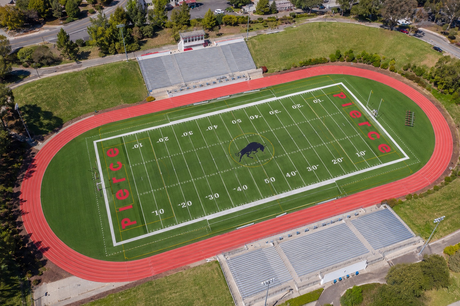 An aerial view of the Brahma Bull stadium at Los Angeles Pierce College, the red running track ringing a green field marked PIERCE in each end zone with the bull at the centre circle, grandstands along both sides.