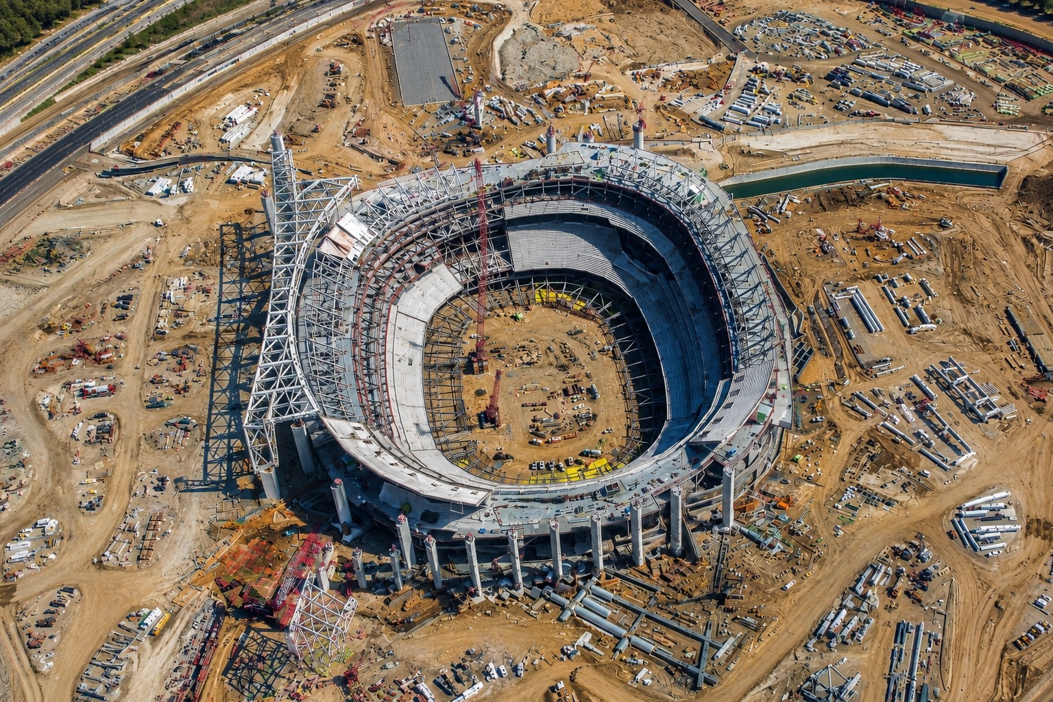 An aerial view of SoFi Stadium under construction, the oval bowl and its canopy steel rising out of a graded site ringed by laydown yards, crane pads, pipe stacks and the water channel at the edge of Hollywood Park.