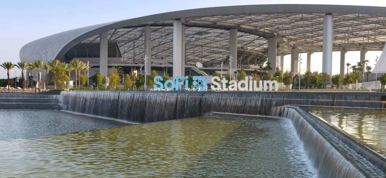 The water feature at SoFi Stadium, a wide cascade falling into the lake in front of the stadium colonnade and the SoFi Stadium letters.