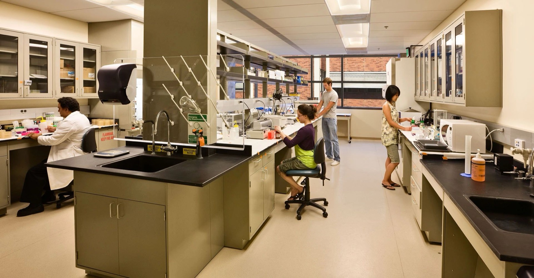 A university research laboratory with fitted casework, fume protection screens and overhead lighting, with researchers at the benches.